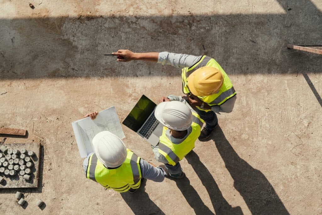 Three construction workers wearing high-visibility vests and hard hats reviewing plans on-site, with one holding blueprints, another using a laptop, and one pointing ahead.