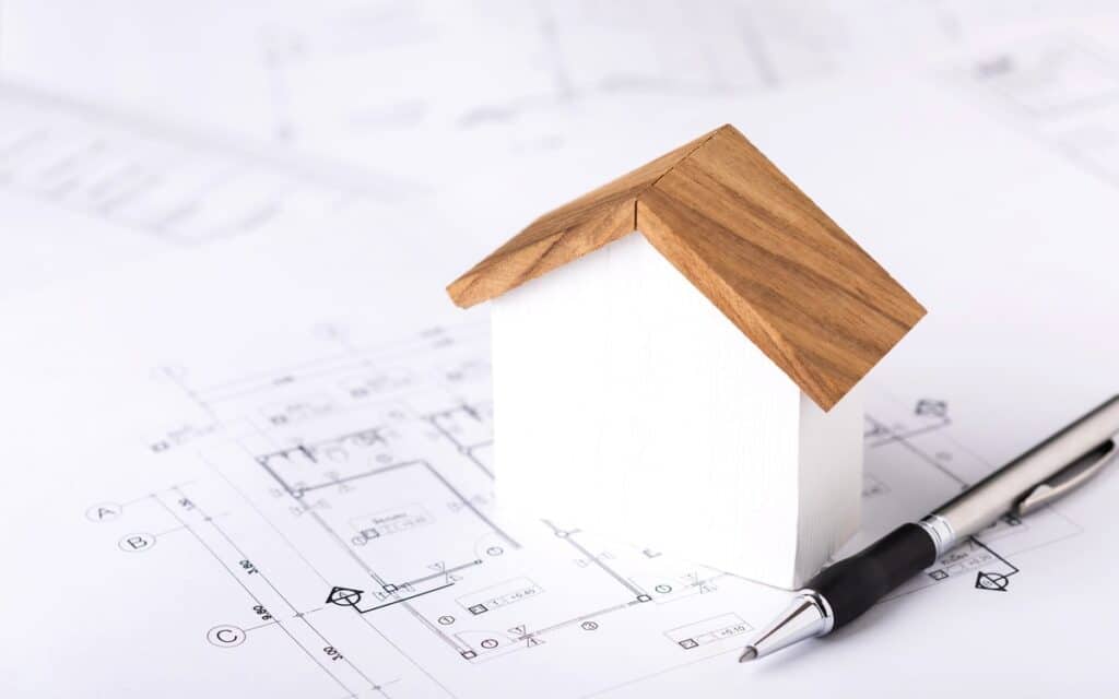 Small wooden house model placed on architectural floor plans with a pen beside it, symbolising home design, planning, or construction documentation.