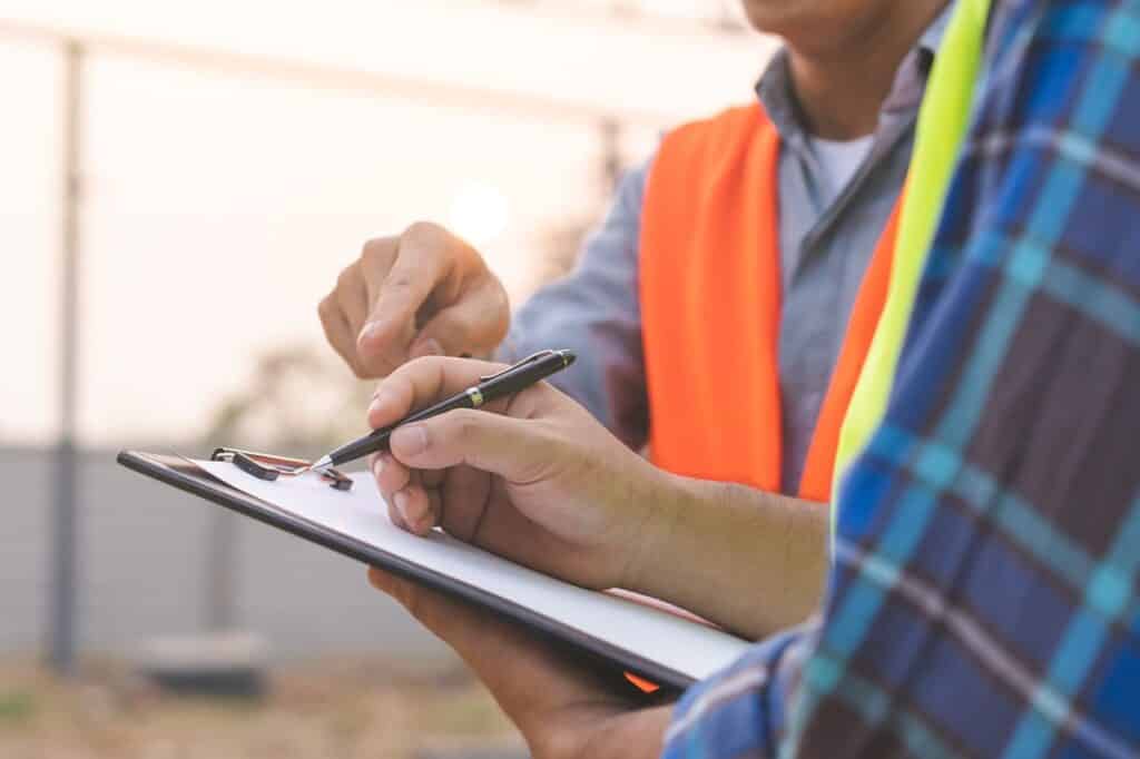 Close-up of two construction workers reviewing a checklist on a clipboard, with one holding a pen and the other pointing, both wearing high-visibility vests.