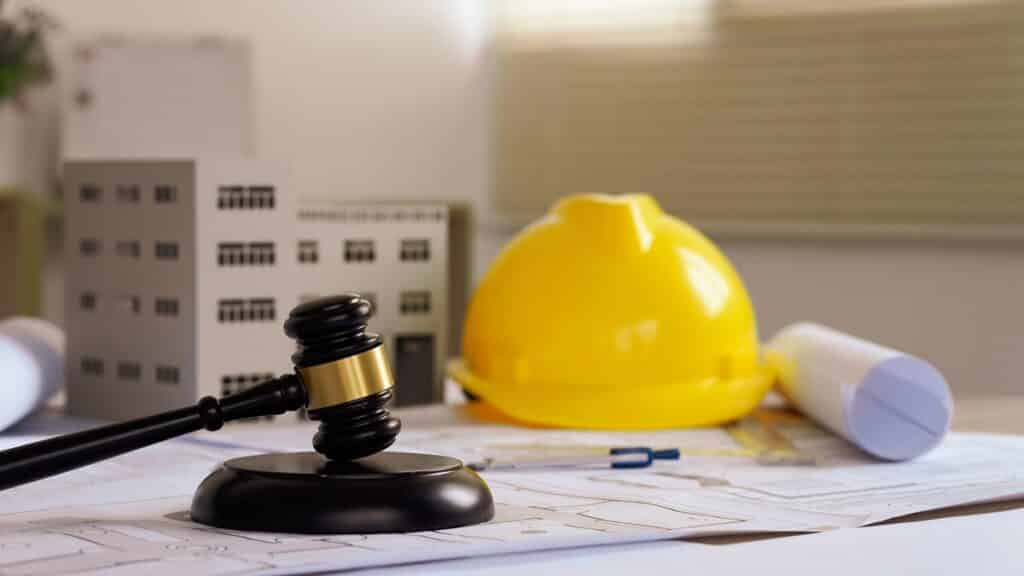 Judge’s gavel resting on architectural plans beside a yellow construction helmet, scale model building, and rolled blueprints, symbolising building laws and regulations.