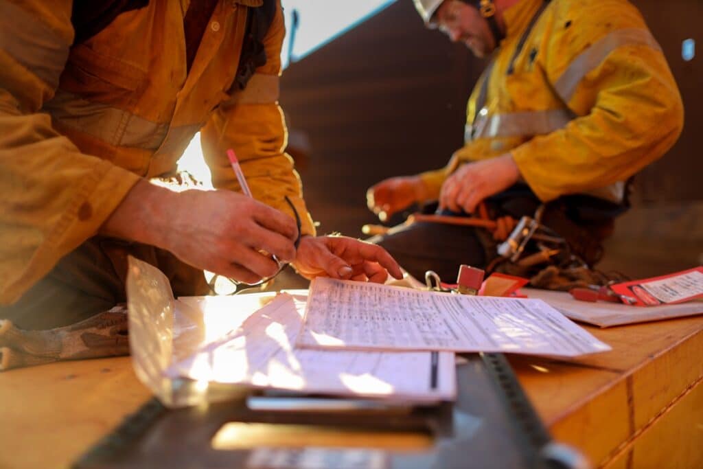 Close-up of a construction worker filling out paperwork on a job site, with safety forms and documents on a wooden surface, while another worker sits in the background.