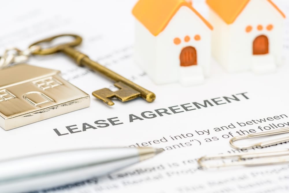 Close-up of a lease agreement document with a gold house key, a house-shaped keyring, miniature house models, and a silver pen placed on top