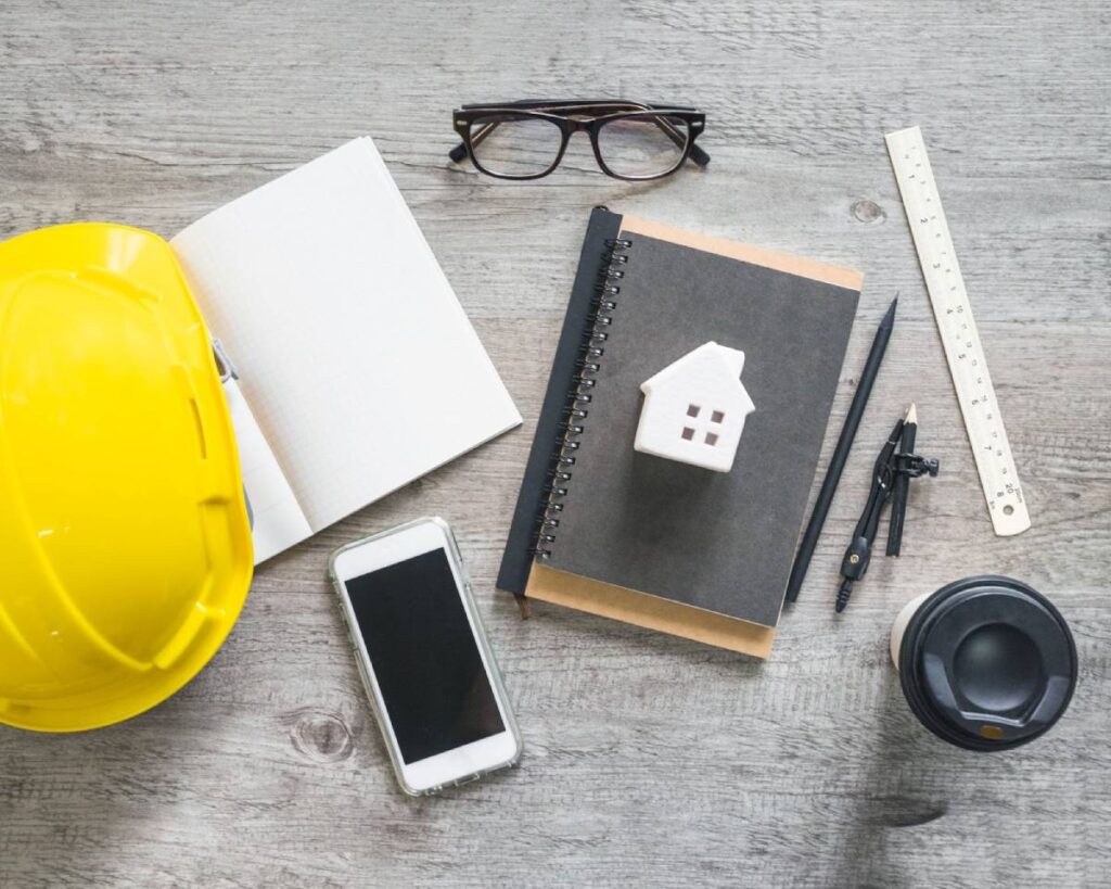 Construction helmet, house model, notebook, and tools on a desk for property planning and inspections.