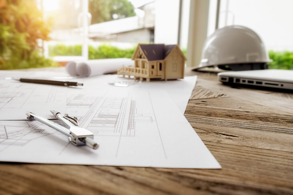 Architectural drawings and drafting tools on a wooden table with a miniature house model, hard hat, and laptop in the background near a bright window.