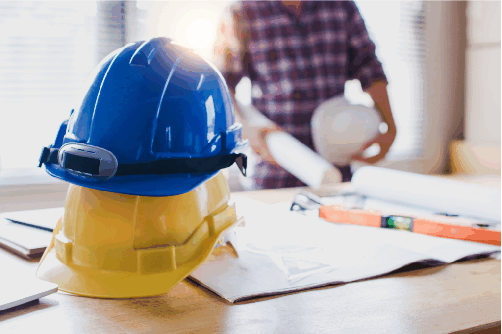 Blue and yellow safety helmets stacked on a desk with rolled-up building plans in the background.