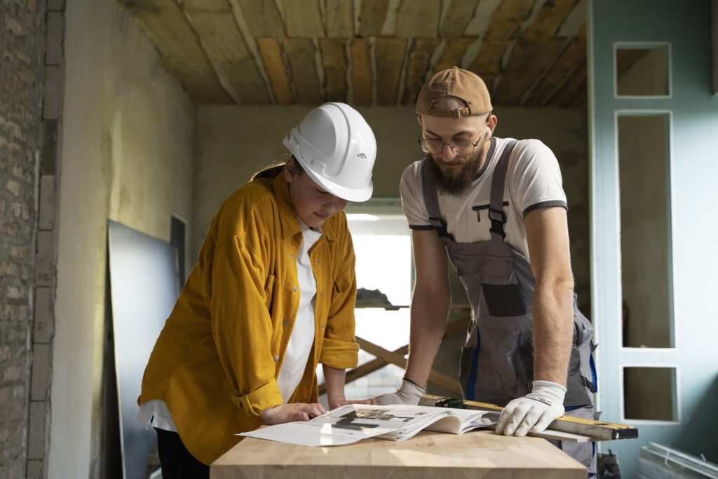 Builder and client reviewing construction plans together inside a partially built home, with timber ceiling and framing visible.