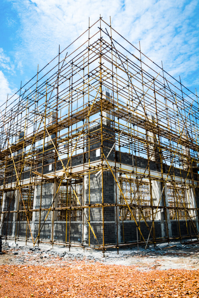 Exterior view of a building under construction, surrounded by full scaffolding against a blue sky, showing the frame and brickwork structure.