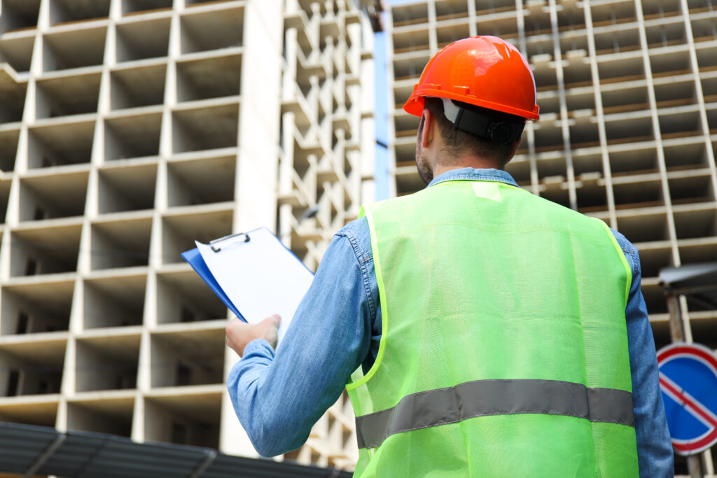 Construction inspector wearing a hard hat and hi-vis vest holding a clipboard while assessing a high-rise building under construction.