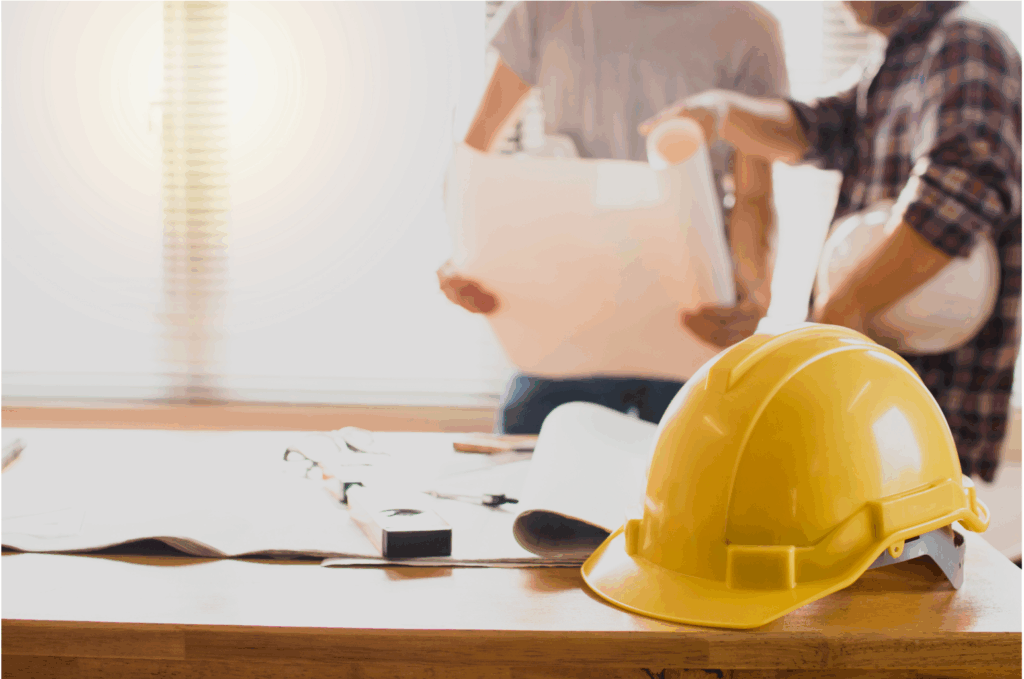 Yellow safety helmet on a desk with construction plans, with two builders discussing blueprints in the background.