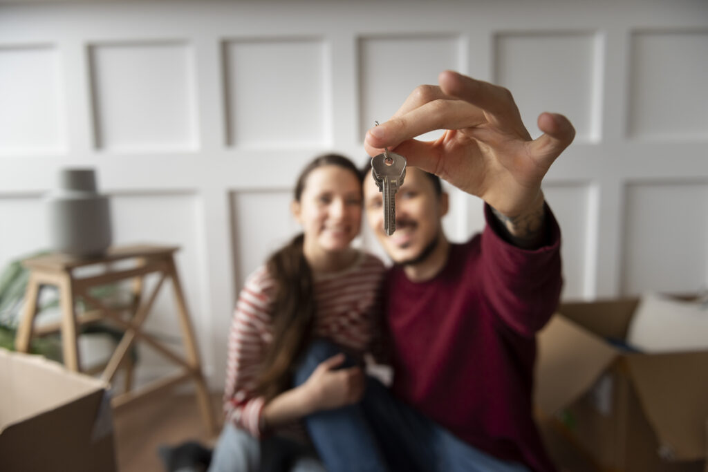 Happy couple sitting in their new home, holding up a set of house keys with moving boxes in the background.