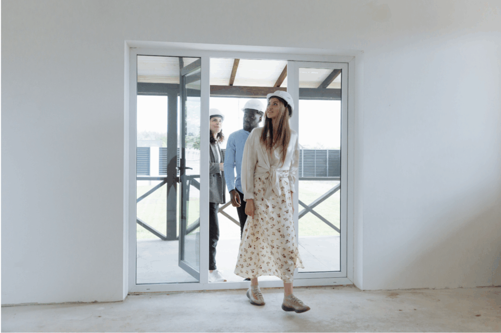 Three people wearing safety helmets inspecting the interior of a new home, entering through large glass sliding doors.
