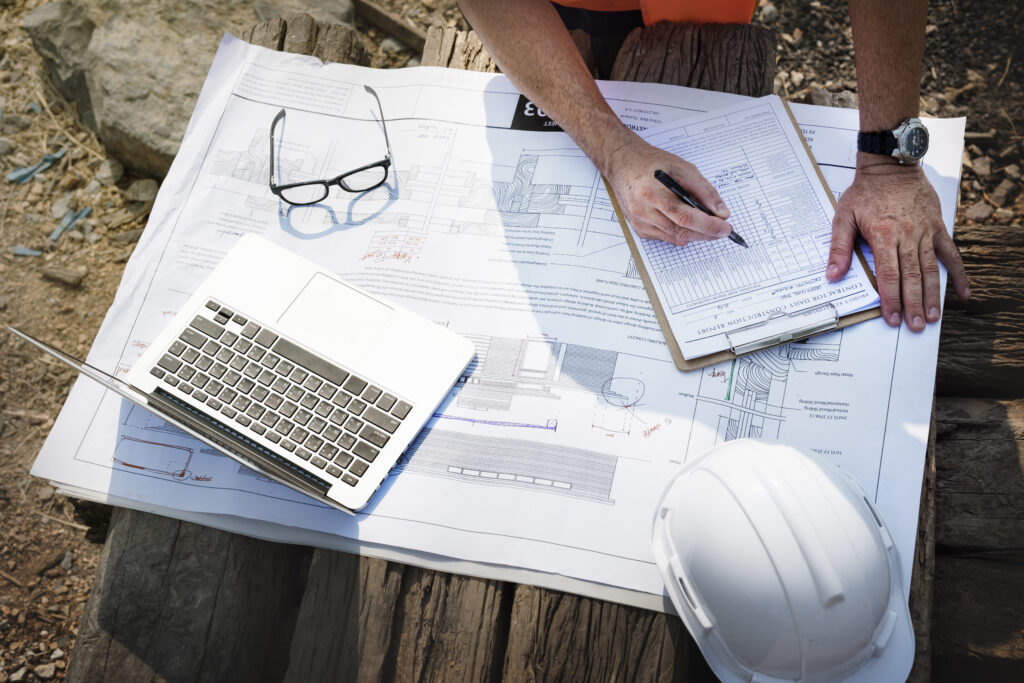 Person reviewing construction plans and filling out a checklist on a clipboard, with a laptop, safety helmet, and glasses on top of technical blueprints.