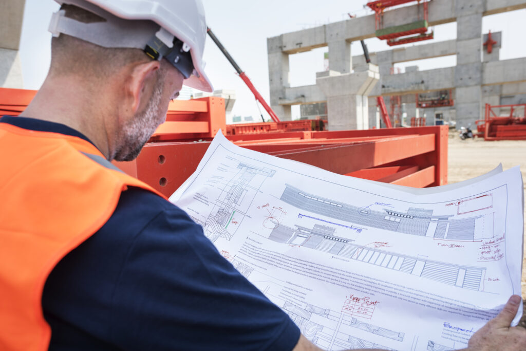 Construction worker in safety gear reviewing architectural plans on site, with red structural components and a partially built concrete framework in the background.