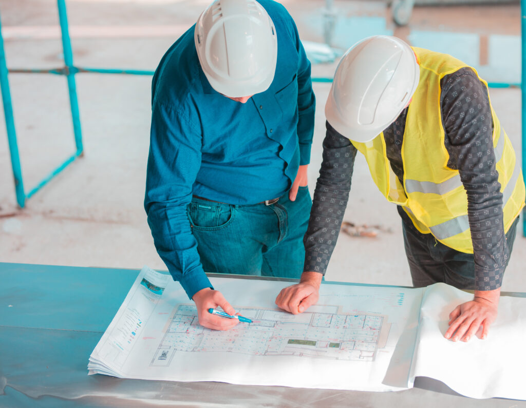 Two home builders leaning over plans, one is holding a pen, and they are both wearing hard hats.