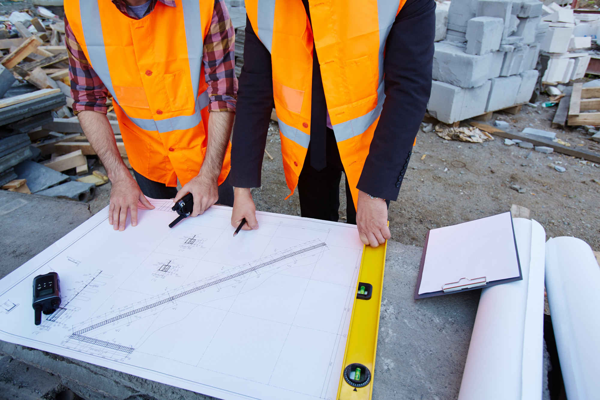 Two construction workers in orange safety vests reviewing detailed building plans on a construction site, with tools, a clipboard, and blueprints nearby.