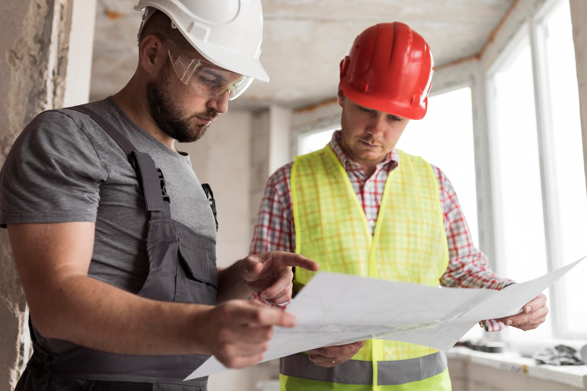 Two construction workers wearing hard hats and safety gear reviewing building plans inside a partially completed structure, discussing next steps on site.