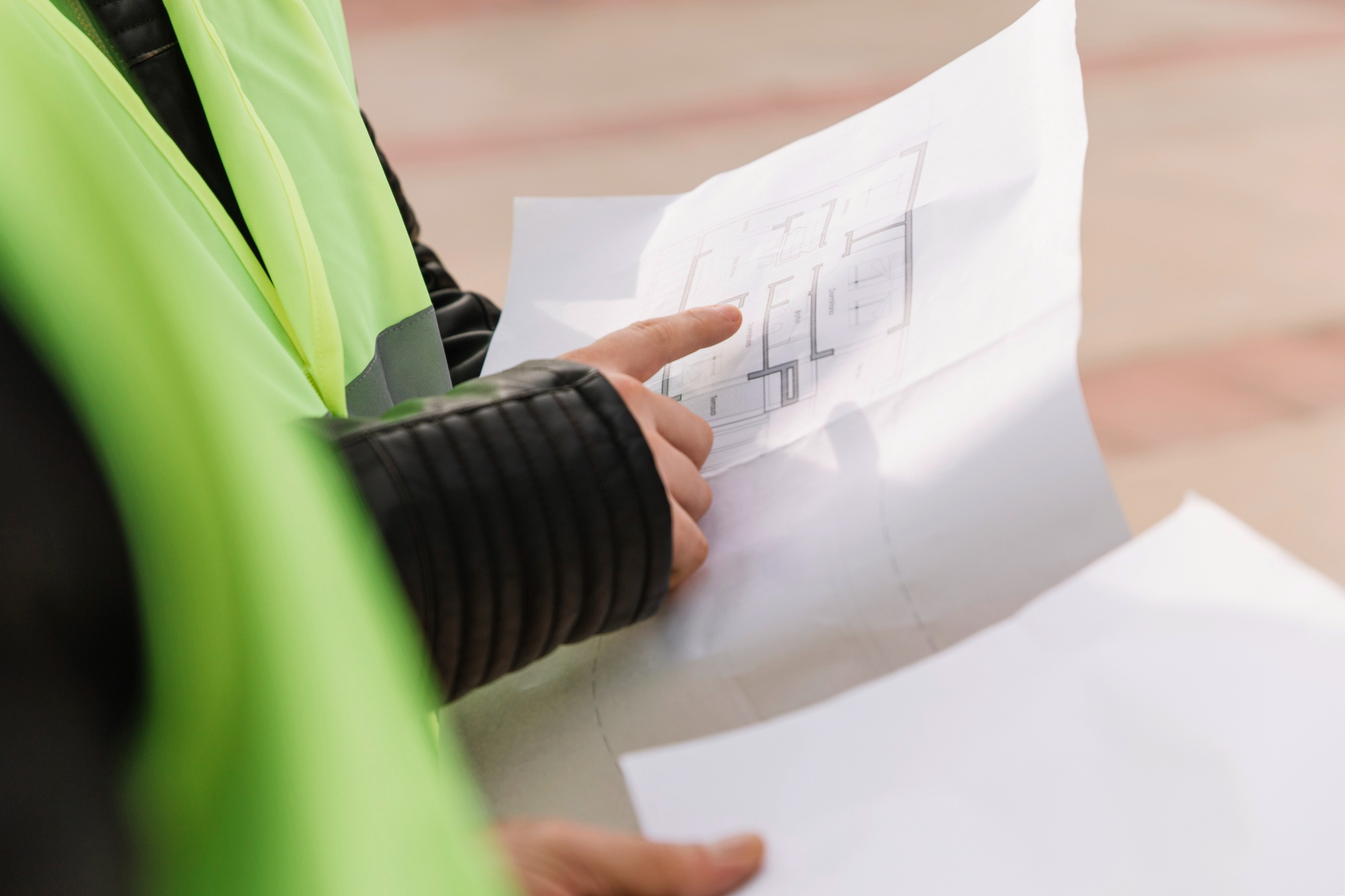 Close-up of a person in a high-visibility vest pointing to a floor plan on a printed architectural drawing during a site check.