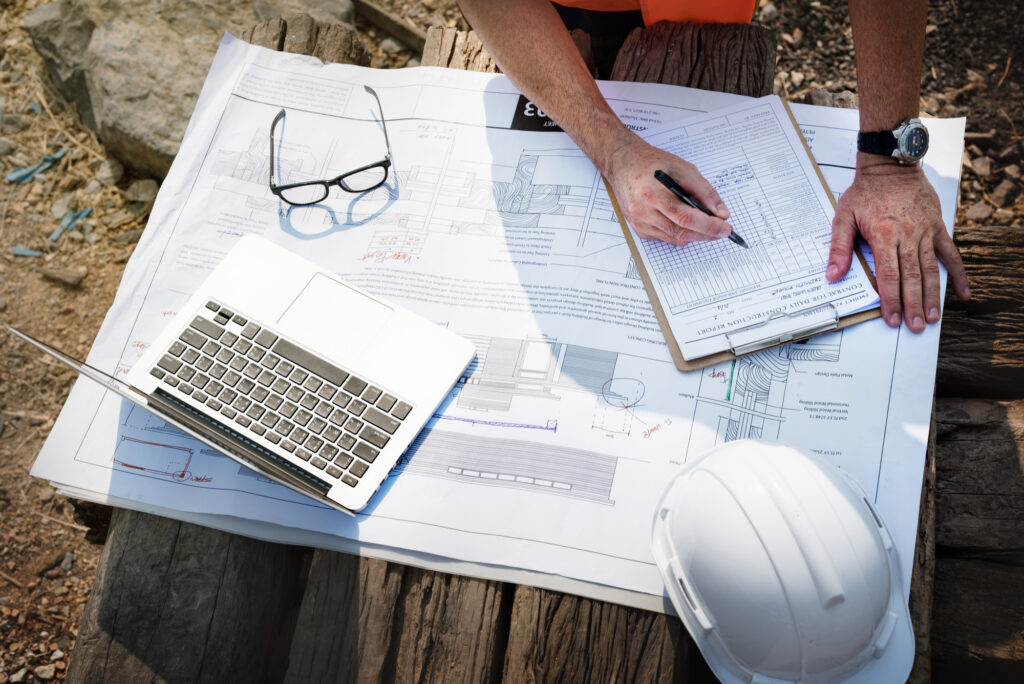 Person completing a checklist on a clipboard over detailed construction plans, with a laptop, hard hat, and glasses on a timber surface at a worksite.