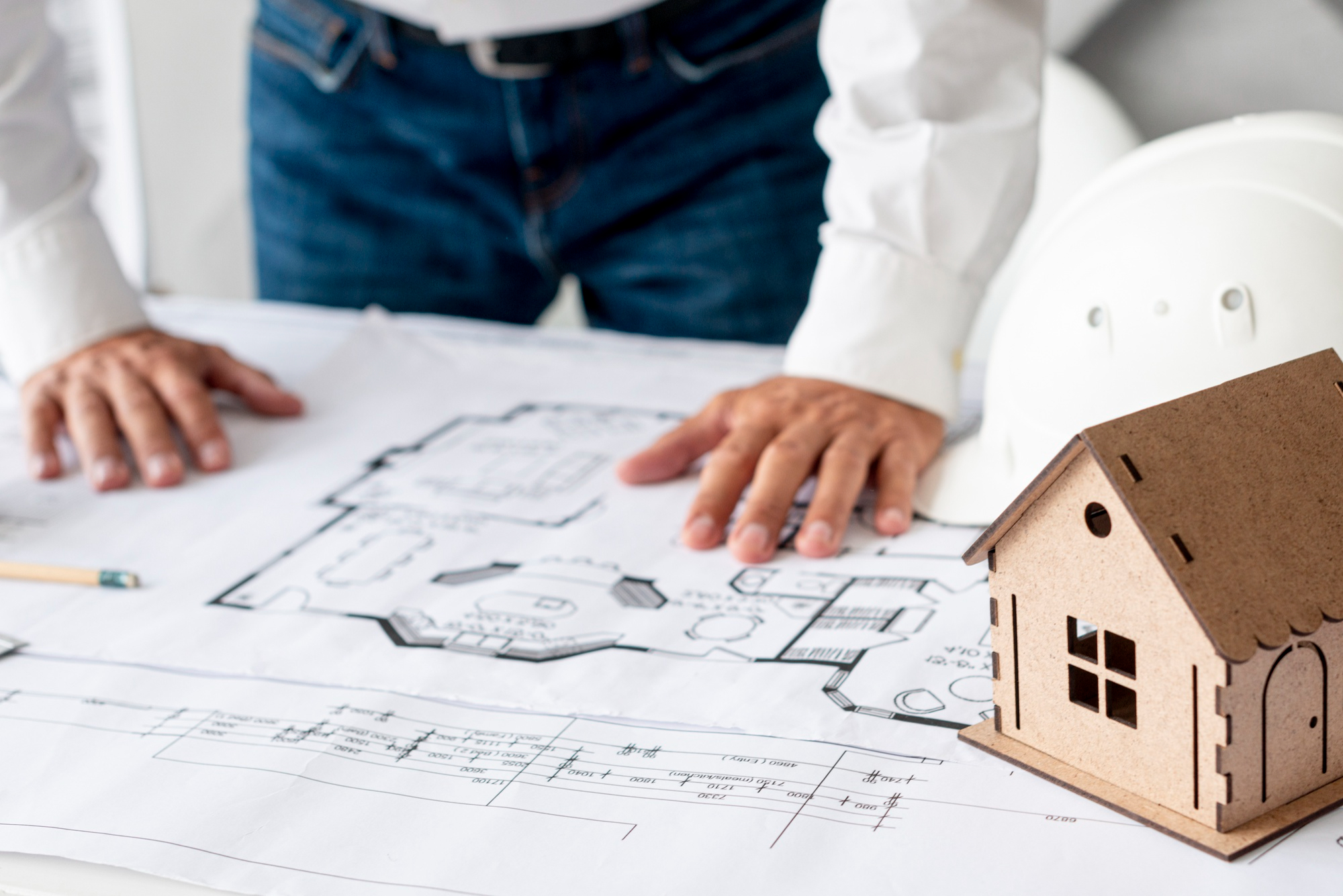 Person standing over architectural blueprints with hands resting on the plans, next to a miniature house model and a white construction helmet.