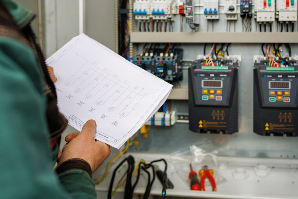 Electrician checking a wiring diagram against an open switchboard during an electrical safety check