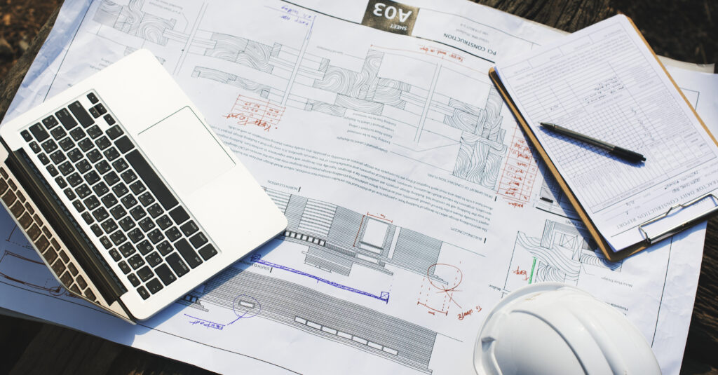 Construction plans spread across a worksite table with a laptop, clipboard, and hard hat.