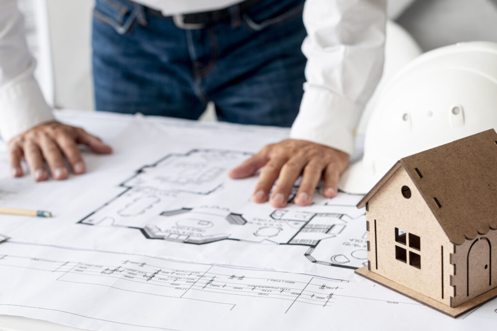 Architect reviewing residential floor plans with a model house and hard hat on a desk.