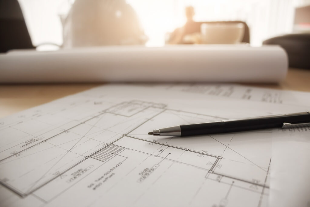 Architectural floor plan on a desk with a drafting pen resting across the building drawings.