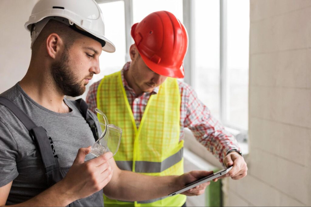 Two workers wearing hard hats and checking notes on a tablet at a residential construction site during the frame stage