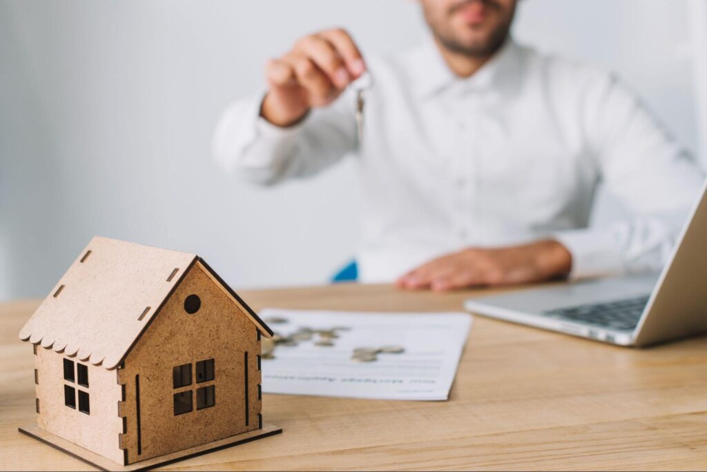 A building inspector holding keys with a wooden house, a contract, and a laptop on the desk in front of him