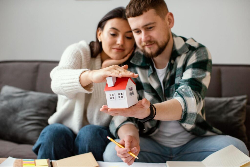 A couple signing paperwork and holding a small house together in their hands.